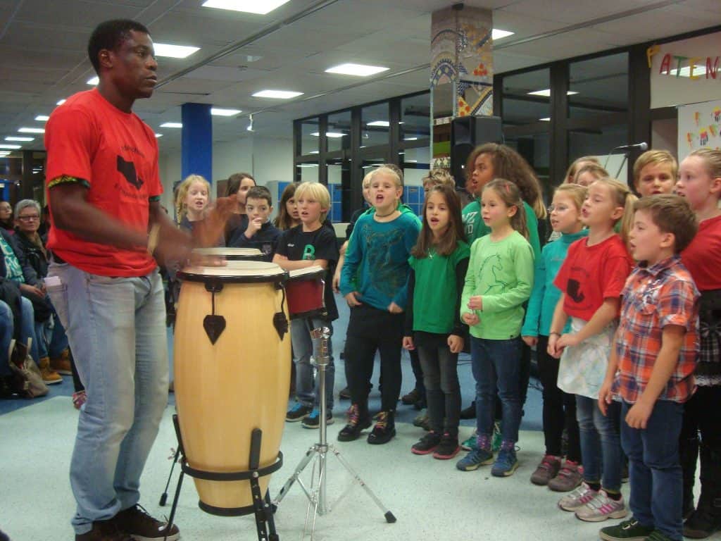 Ein Mann in einem roten Hemd spielt eine große Trommel, während eine Gruppe von Kindern in einem hell erleuchteten Raum in einer Reihe steht, singt oder ihm zusieht. Die Kinder tragen bunte Oberteile, und im Hintergrund sitzen Menschen.