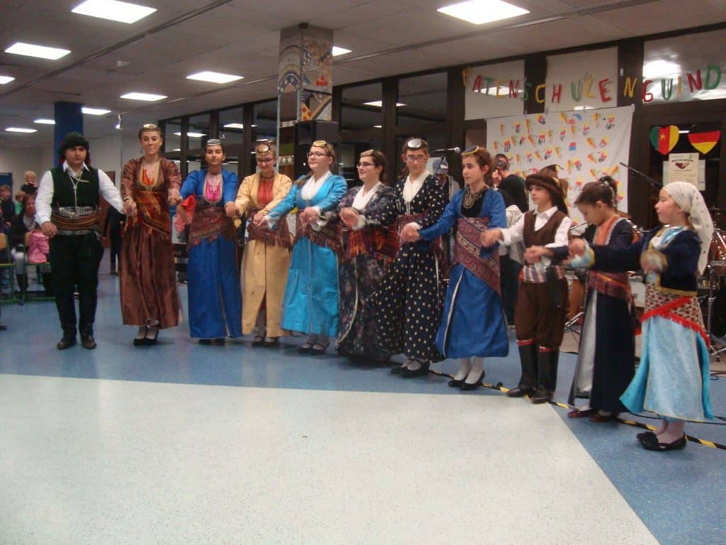 Eine Gruppe von Menschen in traditionellen Trachten steht in einer Reihe und hält sich an den Händen. Es scheint, als würden sie in einem mit farbenfrohen Kunstwerken und Bannern geschmückten Innenraum einen kulturellen Tanz aufführen.