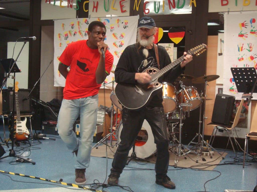 Zwei Musiker treten in einem Raum auf. Einer singt in ein Mikrofon und trägt ein rotes Hemd und Jeans, während der andere in schwarzer Kleidung und mit einer Kappe Gitarre spielt. Im Hintergrund sind Trommeln und andere Instrumente zu sehen.
