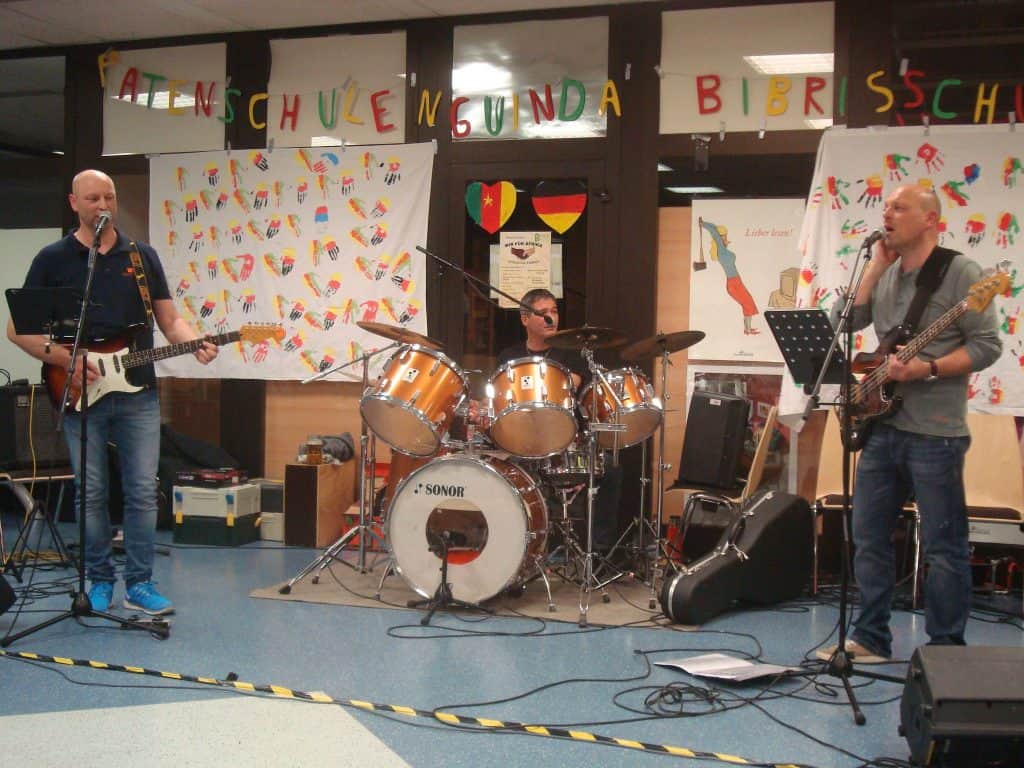 Drei Musiker treten im Innenbereich auf; einer spielt Gitarre und singt, ein anderer Schlagzeug und ein dritter Bass. An der Wand hinter ihnen sind farbenfrohe Kinderkunstwerke und ein Banner zu sehen.