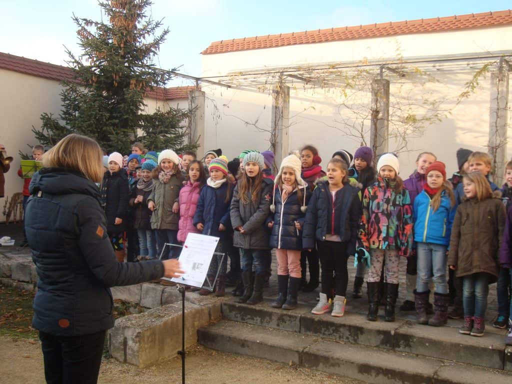 Eine Gruppe winterlich gekleideter Kinder singt draußen auf Steinstufen unter der Leitung eines Erwachsenen mit Notenblättern. Dahinter stehen ein großer immergrüner Baum und eine Pergola.