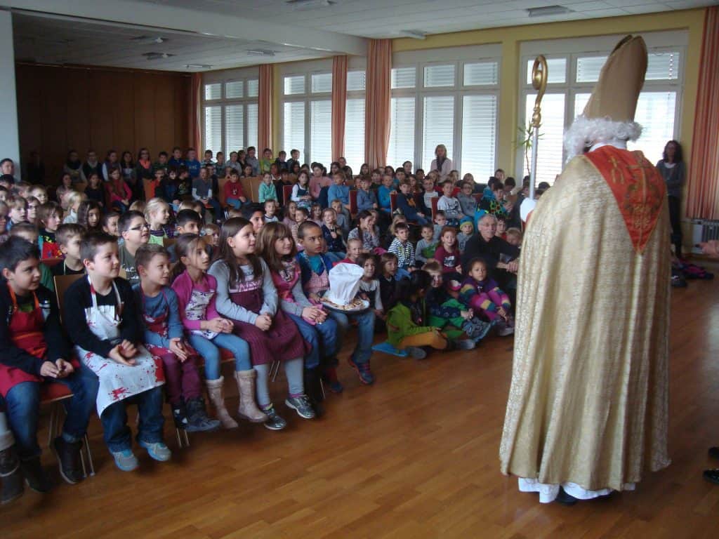 Eine als Nikolaus verkleidete Person steht einer großen Gruppe kleiner Kinder gegenüber, die in Reihen sitzen. Im hinteren Teil eines hellen, geräumigen Raumes sitzen und stehen weitere Kinder und einige Erwachsene.