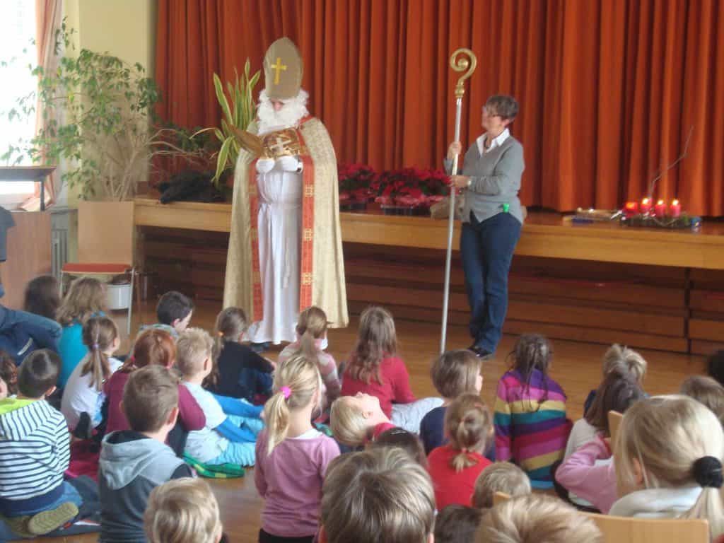 Eine als Sankt Nikolaus verkleidete Person steht neben einer Frau mit einem Stab in der Hand und spricht zu einer Gruppe sitzender Kinder in einer Schulturnhalle mit roten Vorhängen und Weihnachtsdekorationen.