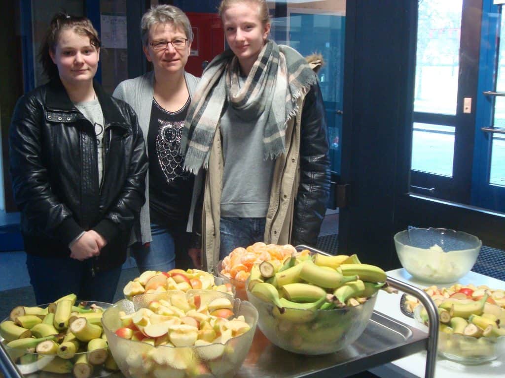 Drei Personen stehen im Innenbereich hinter einem Tisch voller Schalen mit geschnittenen Bananen, Äpfeln und geschälten Mandarinen. Sie tragen Jacken und Schals und scheinen auf einer Veranstaltung oder einem Treffen mit frischem Obst zu sein.