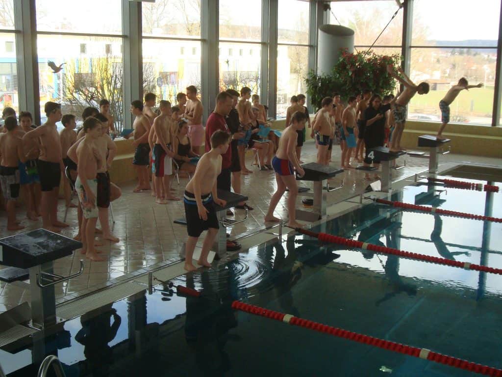 Eine Gruppe Kinder in Badebekleidung steht an einem Hallenbad. Einige bereiten sich auf den Sprung aus den Startblöcken vor, andere warten am Beckenrand. Ein Erwachsener beaufsichtigt die Kinder. Durch große Fenster fällt Sonnenlicht herein.