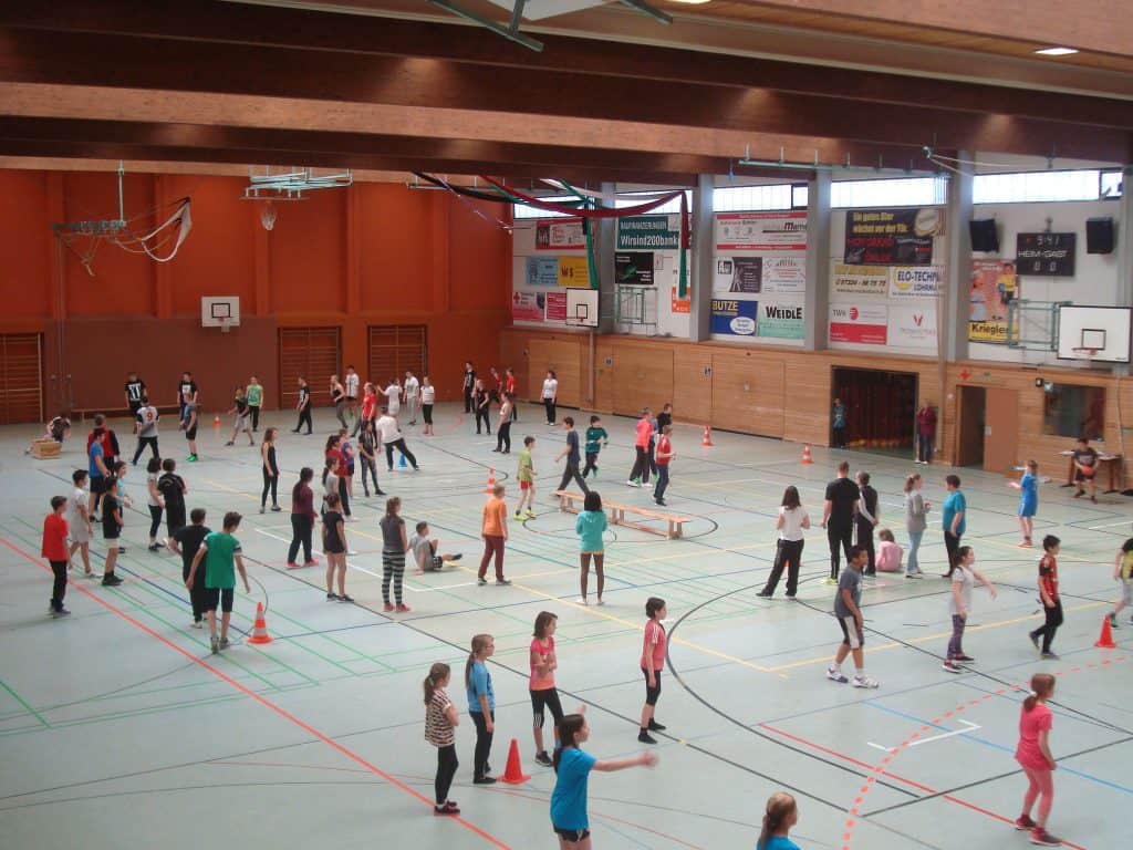 Eine große Gruppe von Kindern und Erwachsenen nimmt an verschiedenen Sportarten und Aktivitäten in einer geräumigen Turnhalle mit Basketballkörben, heller Beleuchtung und Werbebannern an den Wänden teil.