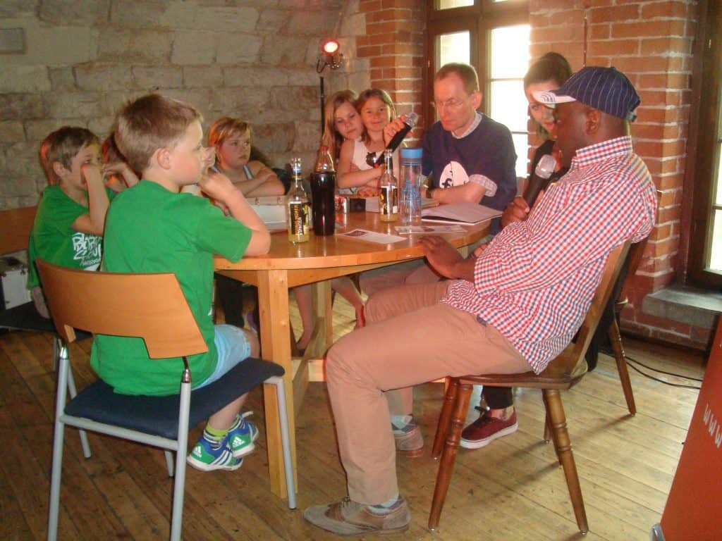 Ein Mann mit einem Mikrofon sitzt mit mehreren Kindern und einem Erwachsenen an einem Tisch. Alle unterhalten sich in einem gemütlichen Raum mit Ziegel- und Steinwänden und Holzboden. Getränke und Bücher stehen auf dem Tisch.