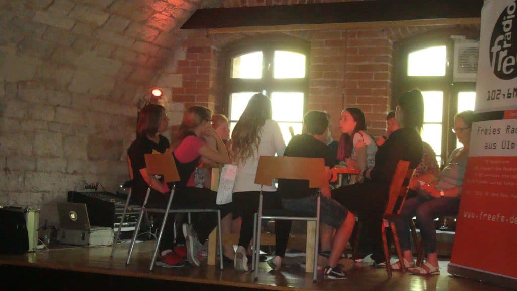 Eine Gruppe von Menschen sitzt im Kreis auf Holzstühlen in einem Raum mit Backsteinwänden und großen Fenstern. Sie scheinen in eine Diskussion vertieft zu sein. Rechts ist ein Banner mit deutschem Text und einem Radio zu sehen.