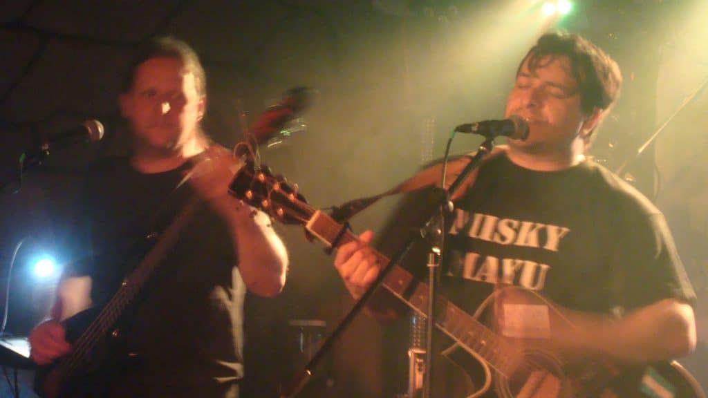 Zwei Musiker treten unter warmem Licht auf der Bühne auf. Einer spielt Bassgitarre, während der andere, ein PUSKY MAYU-Shirt tragend, in ein Mikrofon singt und Akustikgitarre spielt. Der Hintergrund ist verschwommen, Bühnenlichter sind sichtbar.