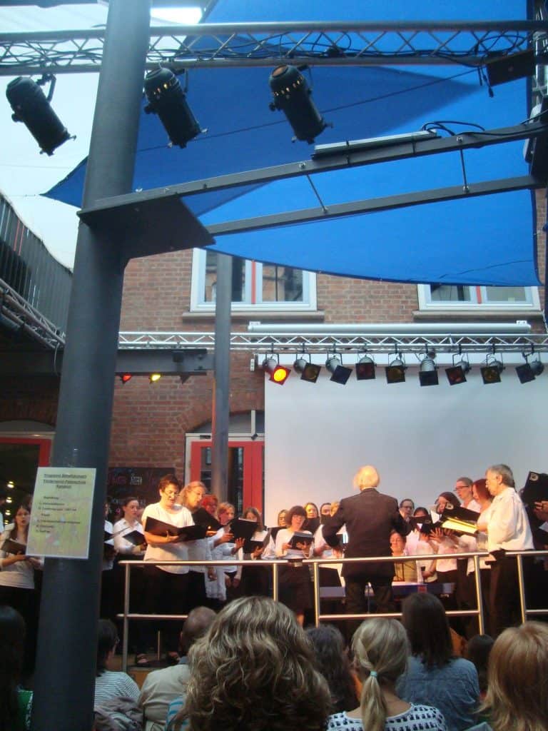 Ein Chor tritt unter blauen Baldachinen auf einer Freilichtbühne auf und wird von einem Dirigenten dirigiert. Im Vordergrund sitzen Zuschauer und verfolgen die Aufführung. Darüber ist eine Beleuchtungsanlage angebracht, im Hintergrund ist ein Backsteingebäude zu sehen.