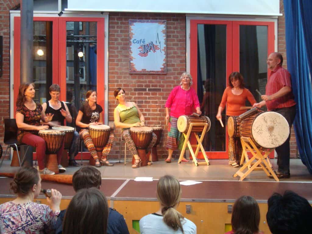 Eine Gruppe von sechs Erwachsenen spielt Schlagzeug auf einer kleinen Bühne, während das Publikum zusieht. Der Hintergrund ist aus Backstein mit großen Fenstern und einem Café-Schild an der Wand. Die Darsteller wirken engagiert und voller Energie.
