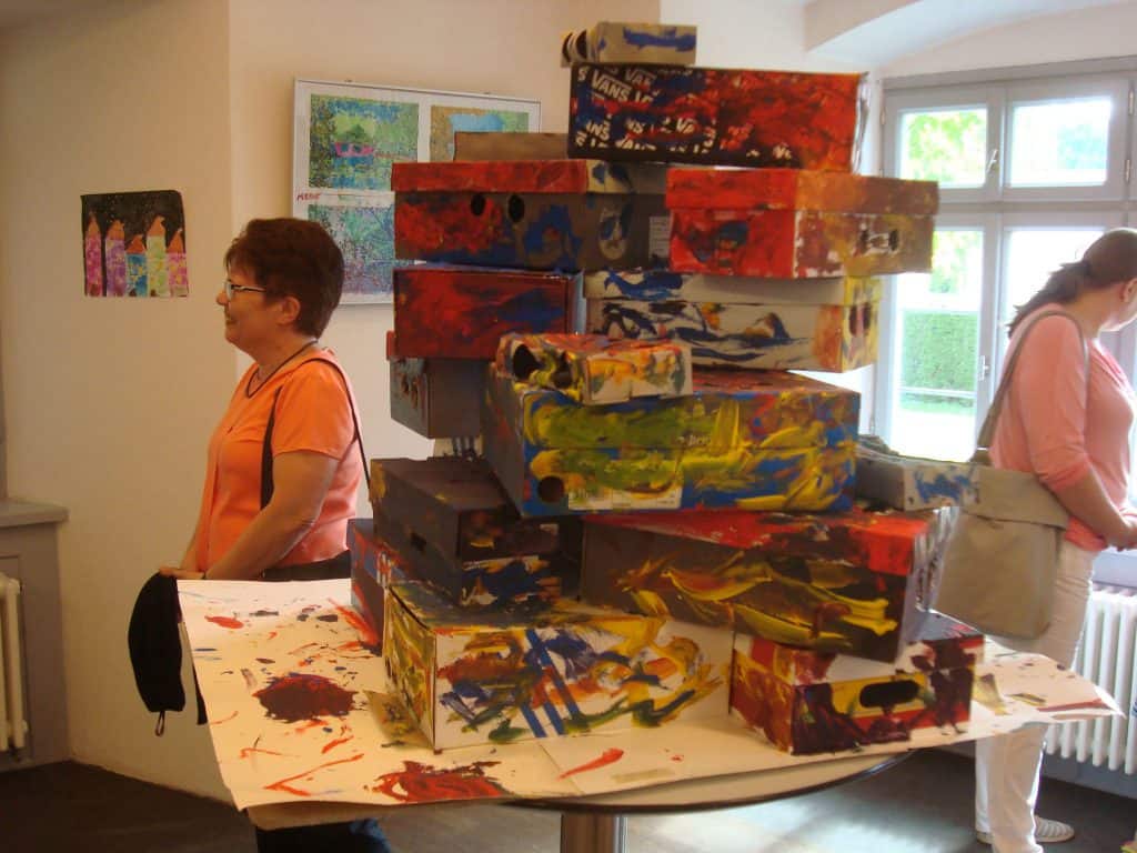 Ein Stapel bunt bemalter Pappkartons steht auf einem Tisch in einer Kunstgalerie. Zwei Frauen stehen daneben. Eine steht, die andere betrachtet die Kunstwerke. An den Wänden hinter ihnen hängen Gemälde.