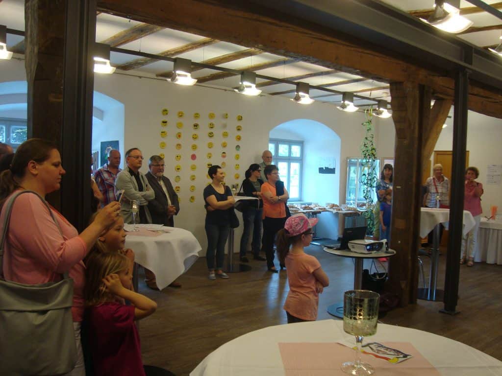 Eine Gruppe von Erwachsenen und Kindern steht in einem hellen Raum mit Holzbalken und hört aufmerksam jemandem zu, der sich außerhalb des Bildes befindet. Überall im Raum stehen weiß gedeckte Tische und Erfrischungen bereit.