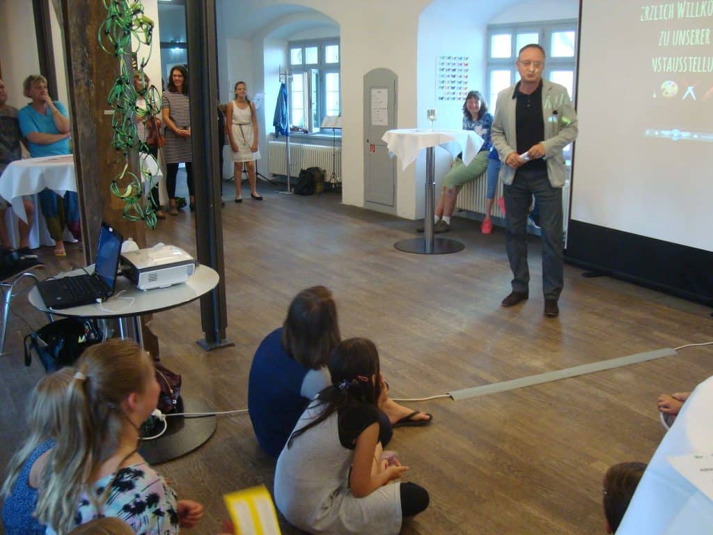 Ein Mann steht vor einer Projektionsleinwand und spricht zu einem kleinen Publikum in einem Raum, während andere stehen und zuschauen. Tische mit Getränken und einem Laptop sind sichtbar und schaffen eine informelle Atmosphäre.