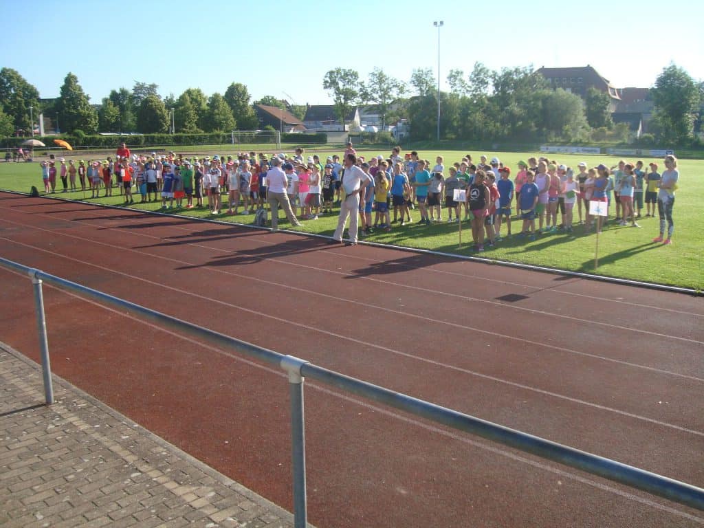 Eine große Gruppe von Kindern und Erwachsenen steht auf einer Wiese neben einer Laufbahn und bereitet sich möglicherweise auf eine Sportveranstaltung an einem sonnigen Tag vor. Im Hintergrund sind Bäume und Gebäude zu sehen.