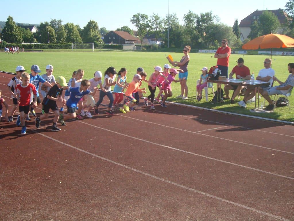 Eine Gruppe Kinder in bunter Sportkleidung startet an einem sonnigen Tag einen Wettlauf auf einer Laufbahn, während Erwachsene und Funktionäre von einem Tisch in der Nähe unter einem orangefarbenen Sonnenschirm aus zuschauen.