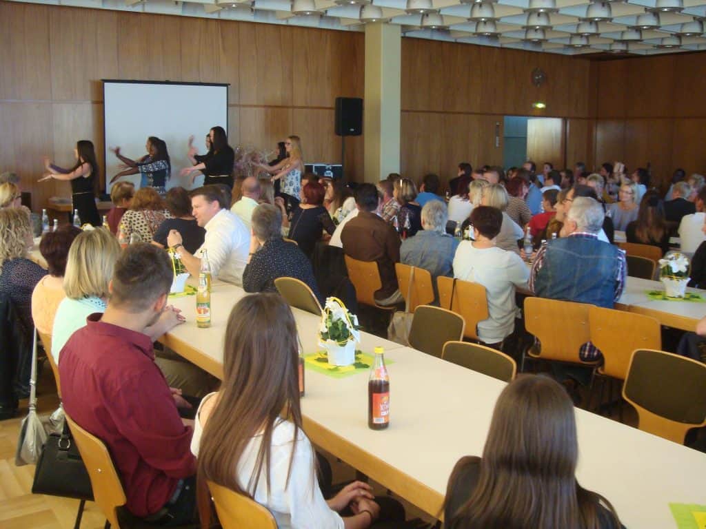 Menschen sitzen an langen Tischen in einem Saal und schauen einer Gruppe von Frauen zu, die vor einer Projektionsleinwand auf der Bühne einen Tanz aufführen. Auf den Tischen stehen Getränke und kleine Blumenarrangements.