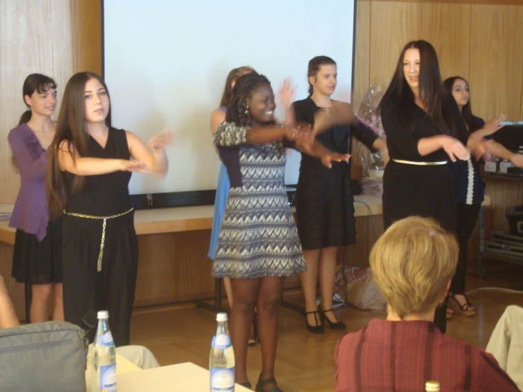 Eine Gruppe junger Frauen steht in einem Klassen- oder Konferenzraum vor einem Bildschirm und führt mit erhobenen Armen einen Tanz auf, während sitzende Personen im Vordergrund zuschauen. Auf dem Tisch stehen Wasserflaschen.