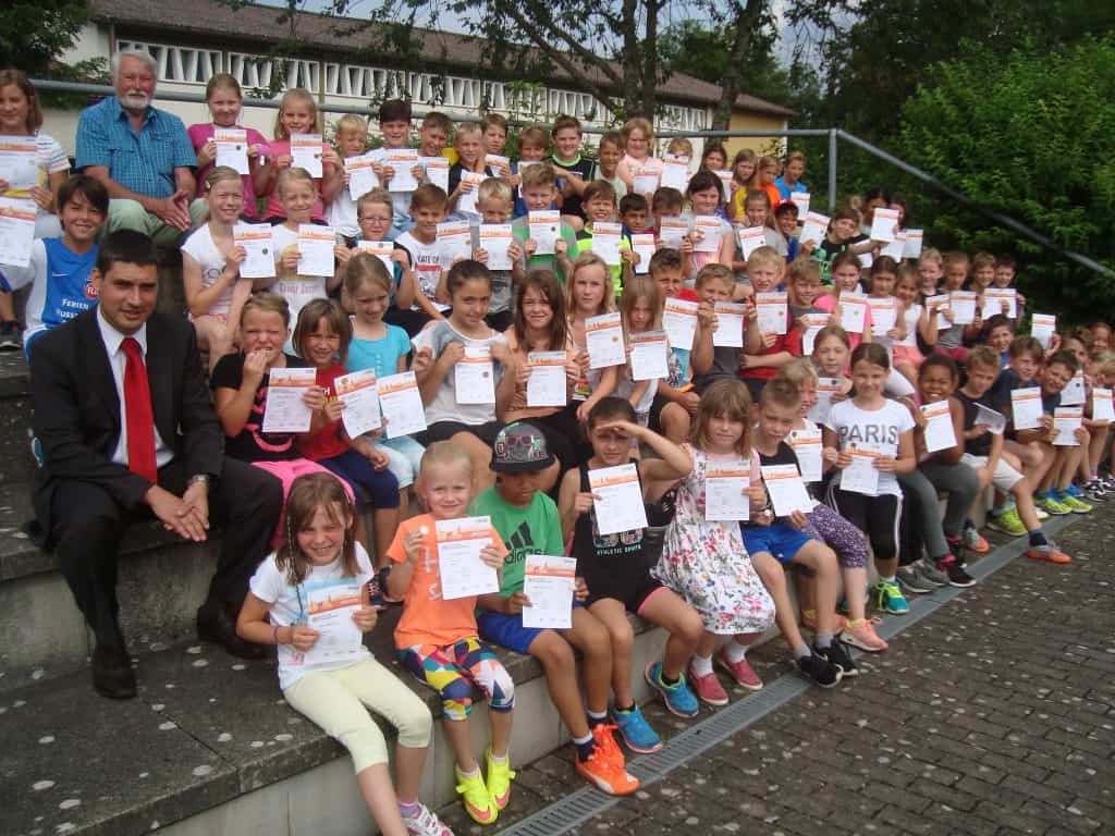 Eine große Gruppe von Kindern und zwei Erwachsenen sitzen lächelnd und mit Zertifikaten in den Händen auf einer Außentreppe, im Hintergrund sind Grünflächen und ein Gebäude zu sehen.