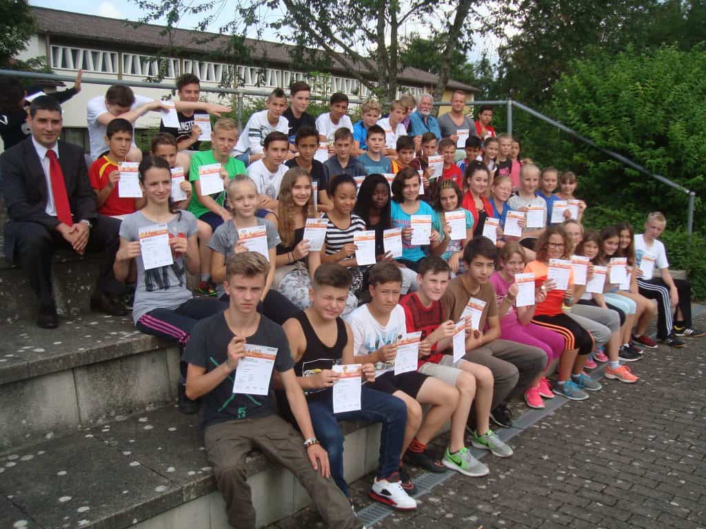 Eine große Gruppe Kinder und ein Mann im Anzug sitzen lächelnd auf einer Außentreppe und halten Zertifikate hoch. Im Hintergrund sind Bäume und ein Gebäude zu sehen.