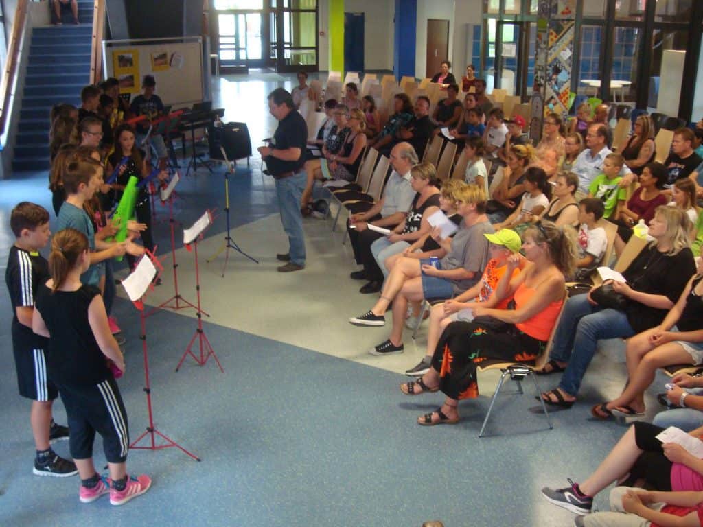 Eine Gruppe von Kindern steht vor Notenständern und spielt für ein in Reihen sitzendes Publikum in einem hellen Innenraum. Ein Erwachsener steht in der Mitte und leitet die Veranstaltung.