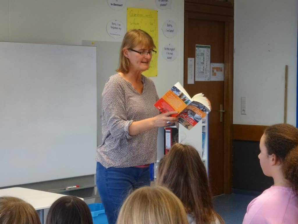 Eine Frau steht vor einem Klassenzimmer. Sie hält ein Buch in der Hand und liest einer Gruppe von Schülern vor, die ihr gegenüber sitzen. An der Wand des Klassenzimmers hängen Plakate, im Hintergrund ist eine geschlossene Tür zu sehen.