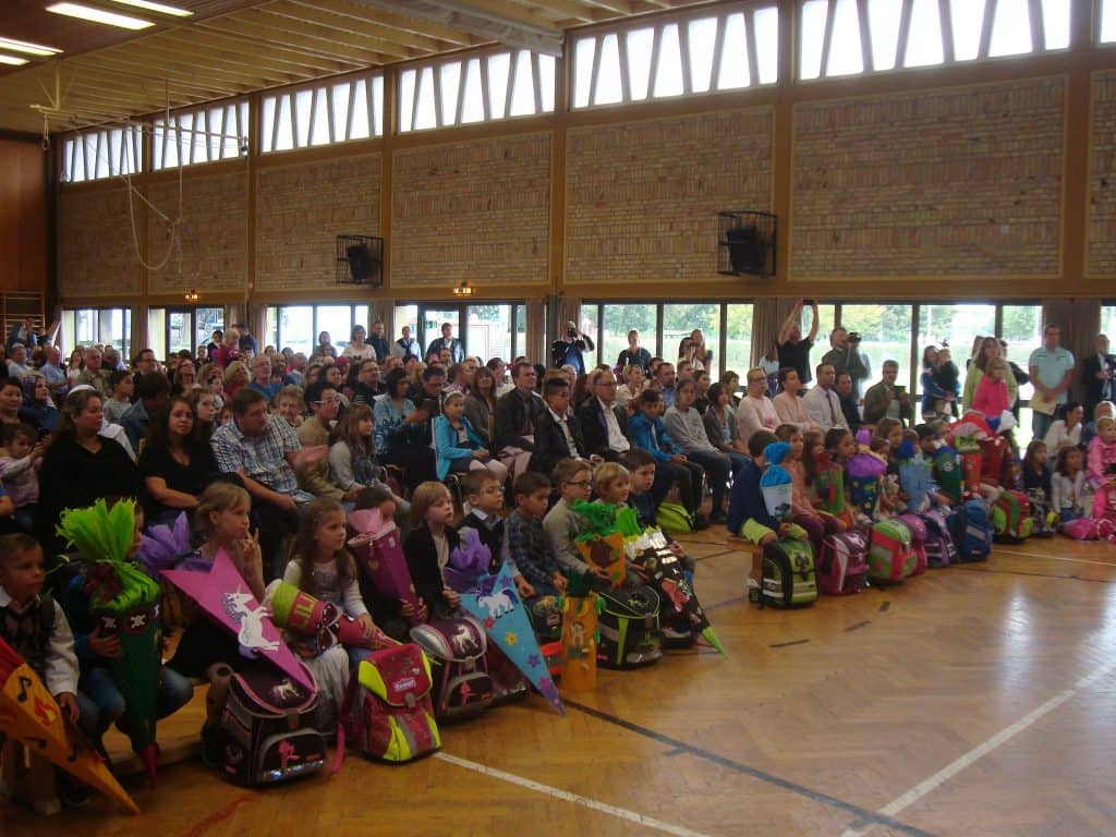 Eine große Gruppe Kinder mit bunten Schultüten sitzt in der ersten Reihe einer Turnhalle, während dahinter Eltern und Erwachsene sitzen und einer Schulveranstaltung oder Feier beiwohnen.