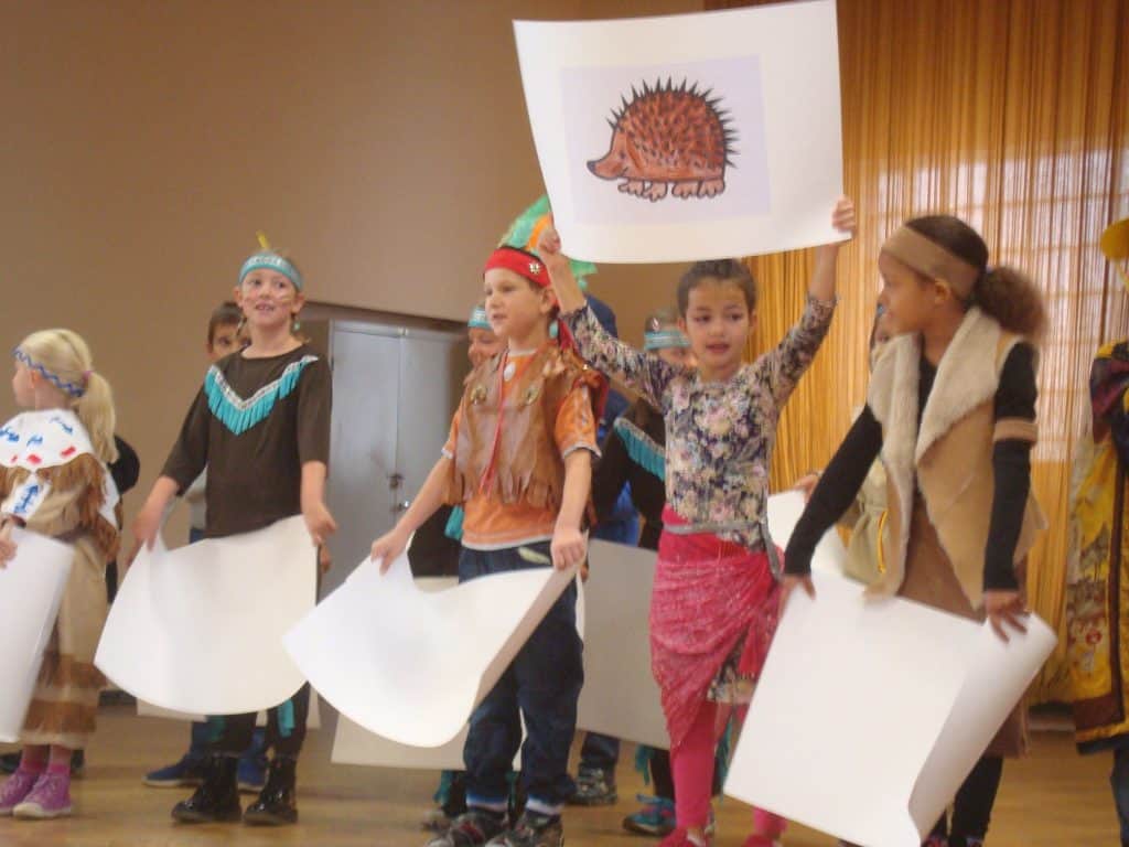 Eine Gruppe kleiner Kinder in Kostümen steht in einer Reihe auf der Bühne. Ein Mädchen hält eine große Zeichnung eines Igels hoch, die anderen halten leere weiße Blätter. Sie scheinen an einem Schultheaterstück oder einer Aufführung teilzunehmen.