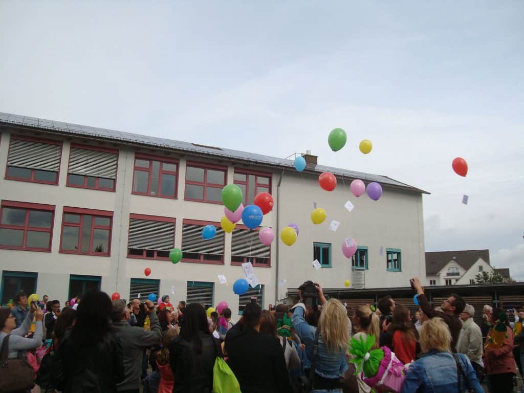 Eine Menschenmenge lässt vor einem Schulgebäude bunte Luftballons in den Himmel steigen und beobachtet, wie sie nach oben schweben. An einigen Ballons sind kleine Zettel befestigt. Es ist ein bewölkter Tag.