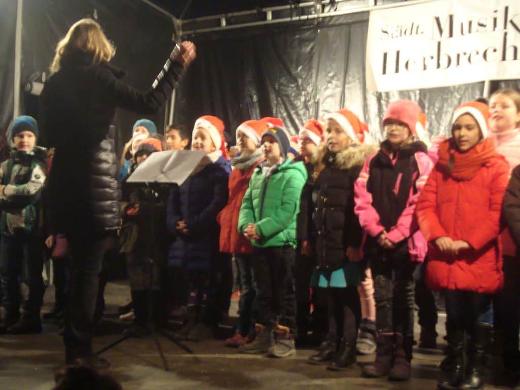 Eine Gruppe Kinder in Wintermänteln und Weihnachtsmannmützen singt auf der Bühne, angeführt von einer Dirigentin. Vor ihnen steht ein Notenständer, im Hintergrund hängt ein Schild.