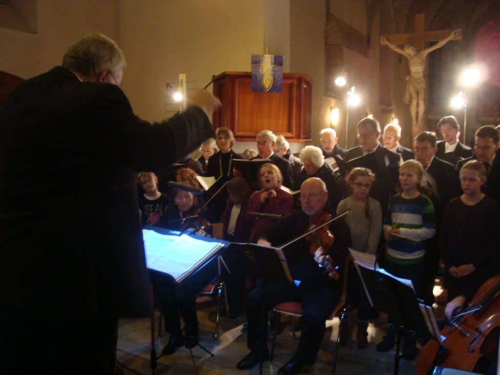 Ein Dirigent leitet einen Chor und ein Orchester, die in einer schwach beleuchteten Kirche auftreten. Sänger und Musiker sitzen und stehen, im Hintergrund ist ein Kruzifix zu sehen.