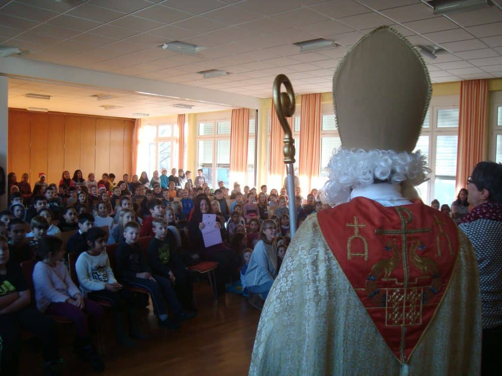 Eine als Bischof verkleidete Person mit Bischofsstab steht in einem hell erleuchteten Raum vor einer Gruppe sitzender Kinder und spricht zum Publikum. Die Kinder schauen aufmerksam zu.