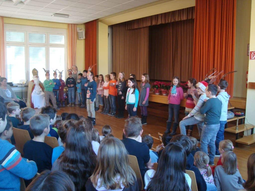 Eine Gruppe von Kindern steht auf der Bühne und tritt vor einem Publikum aus sitzenden Kindern in einer Schulaula auf. Einige Kinder tragen Rentiergeweihe, was auf ein weihnachtliches oder winterliches Theaterstück hindeutet.