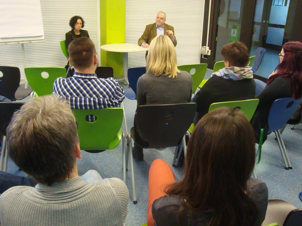 Eine kleine Gruppe von Menschen sitzt auf bunten Stühlen vorn im Raum und sieht zwei Sprechern gegenüber, einer Frau und einem Mann, die an einer Diskussion oder einem Seminar teilnehmen.