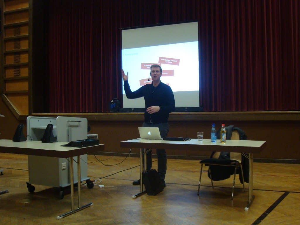 Eine Person steht und gestikuliert, während sie in einem Raum mit roten Vorhängen vor einer Leinwand eine Präsentation hält. Auf dem Tisch vor ihr stehen ein Laptop und eine Wasserflasche.