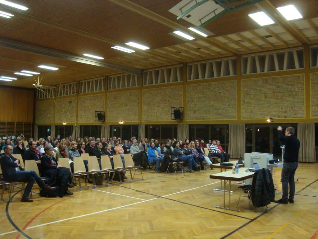 Ein Redner steht vor einem Publikum, das in Stuhlreihen in einer großen Turnhalle mit Holzboden und hohen Decken sitzt. Vorne sind Tische und ein Projektor aufgestellt.