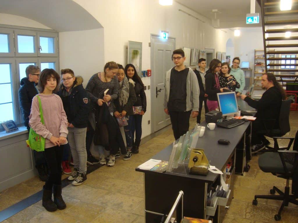 Eine Gruppe überwiegend junger Menschen steht in einer Schlange vor einem Empfangstresen in einem hellen Gebäude. Eine Person sitzt am Schreibtisch und nutzt einen Computer, während die Gruppe wartet und miteinander kommuniziert.