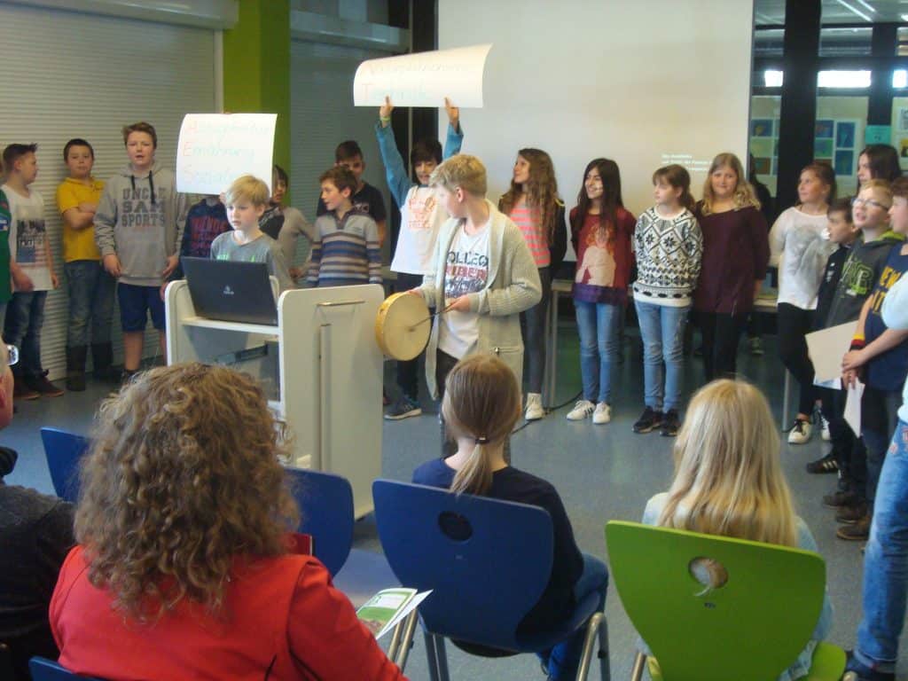 Eine Gruppe von Kindern steht und sitzt in einem Klassenzimmer. Einige halten Schilder hoch, während ein Kind an einem Podium spricht und ein anderes Trommel spielt. Mehrere Personen sitzen davor und verfolgen die Präsentation.
