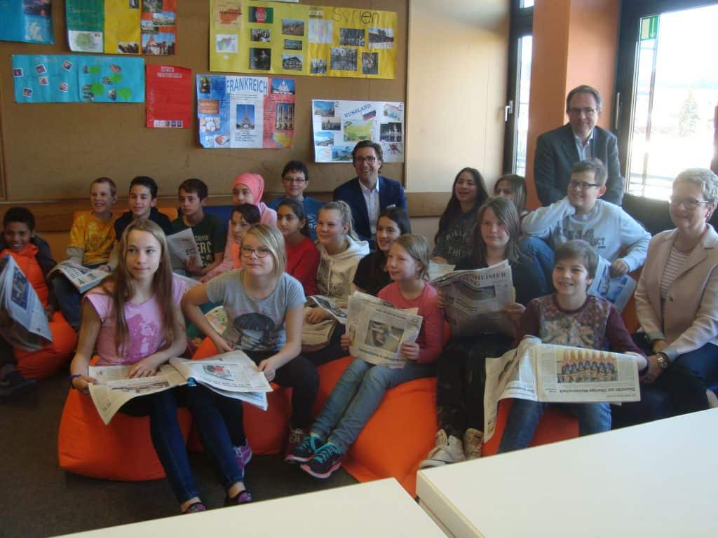 Eine Gruppe von Schülern sitzt mit Zeitungen in den Händen auf orangefarbenen Sitzsäcken in einem Klassenzimmer. Vier Erwachsene stehen und sitzen lächelnd zwischen ihnen. Im Hintergrund sind bunte Plakate und Fenster zu sehen.