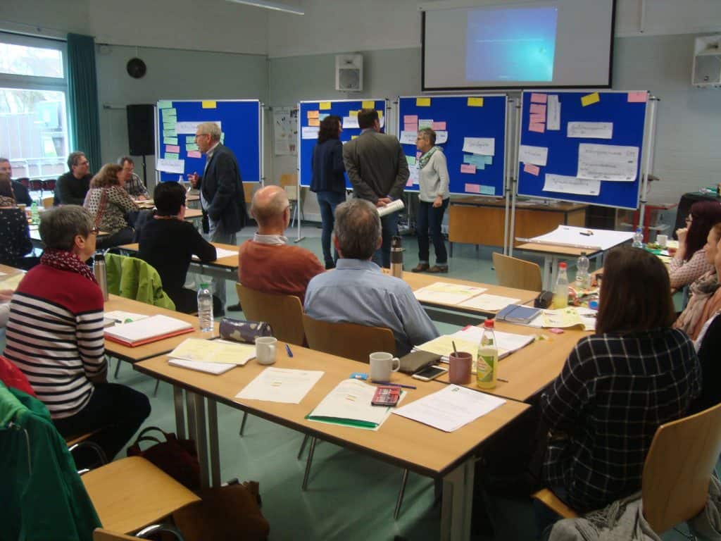 An Tischen sitzende Personen beobachten eine Gruppe, die in einem Klassenzimmer oder einer Werkstatt an blauen Tafeln steht, die mit Notizen und Papieren bedeckt sind. Materialien, Notizbücher und Getränke stehen auf den Tischen, im Hintergrund läuft eine Projektionsleinwand.