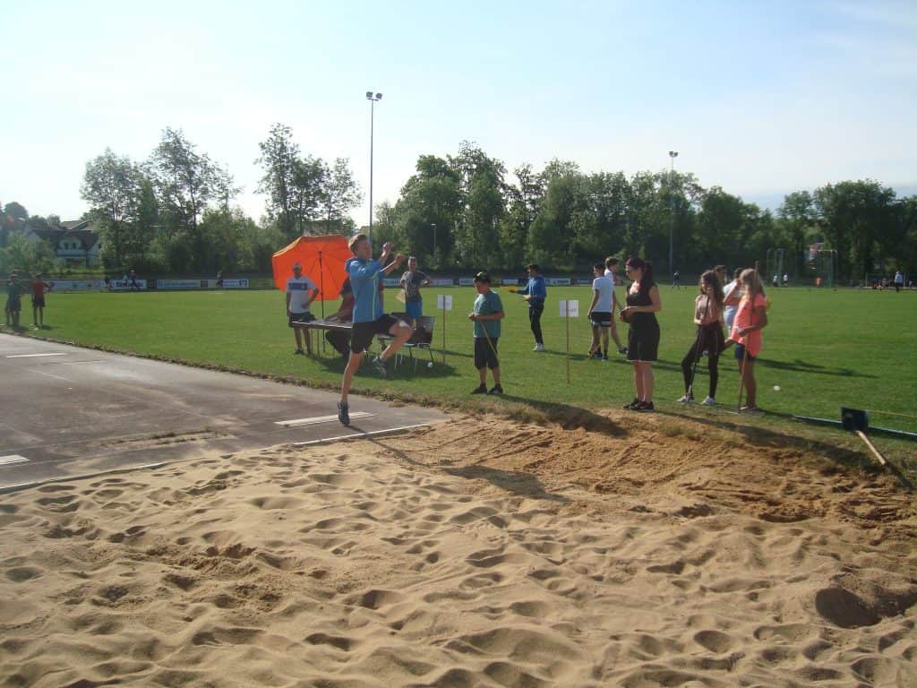 Ein junger Athlet versucht bei einem Laufwettbewerb im Freien in der Luft einen Weitsprung in eine Sandgrube, während Zuschauer und Offizielle an einem sonnigen Tag in der Nähe zuschauen.