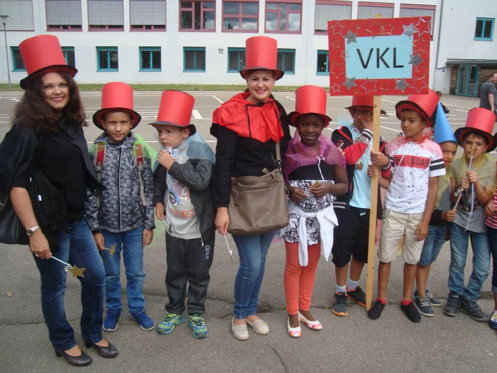 Eine Gruppe von Kindern und zwei Erwachsenen steht vor einem Gebäude. Alle tragen rote Zylinder. Einige halten Spielzeug oder Zauberstäbe in den Händen, ein Erwachsener hält ein VKL-Schild. Sie scheinen an einer festlichen Veranstaltung oder Parade teilzunehmen.