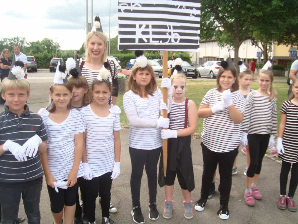 Eine Gruppe von Kindern und eine Frau stehen im Freien. Sie tragen schwarz-weiß gestreifte Hemden, weiße Handschuhe und Kostümhüte mit Bommeln, einige sind mit Gesichtsbemalung versehen. Sie halten ein gestreiftes Schild und scheinen an einer Themenveranstaltung oder Parade teilzunehmen.