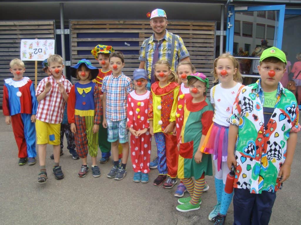 Eine Gruppe lächelnder Kinder und ein Erwachsener, alle in bunten Clownkostümen und mit roten Nasen, stehen gemeinsam im Freien vor einem Gebäude.