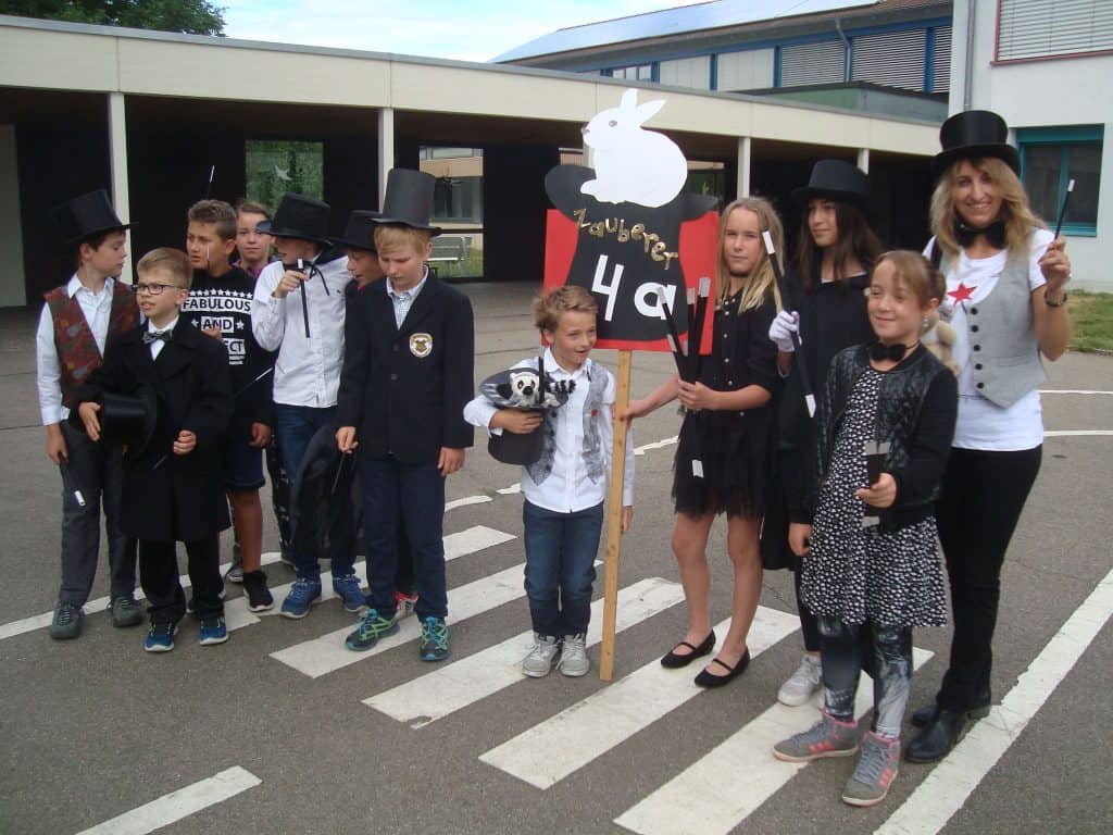 Eine Gruppe von Kindern und ein Erwachsener, die als Zauberer verkleidet sind, einige tragen Zylinder und halten Zauberrequisiten in den Händen, stehen draußen auf dem Bürgersteig. Ein Kind hält ein Zaubererschild mit einem Hasenmotiv.