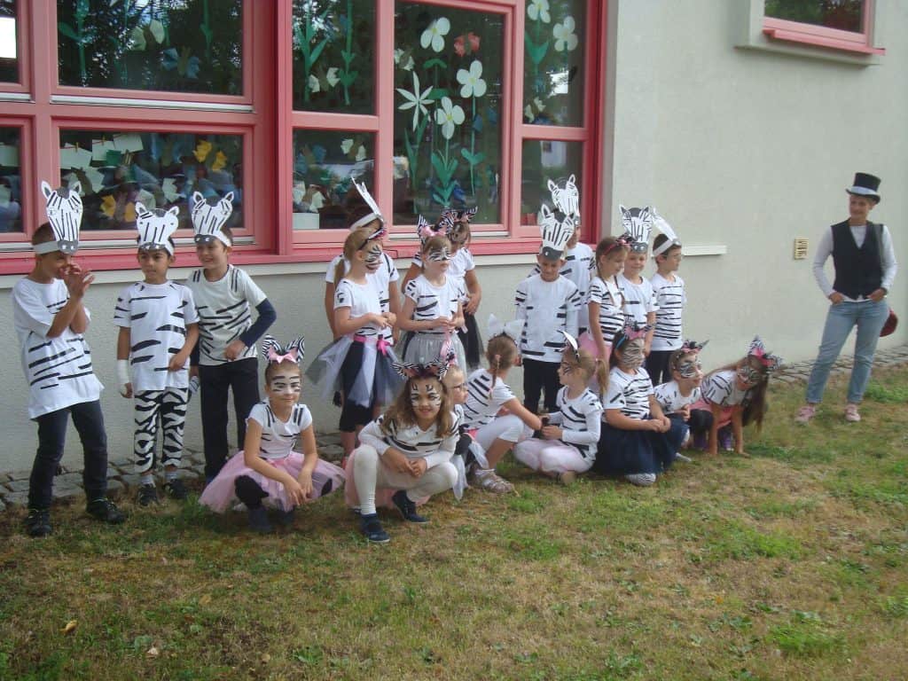 Eine Gruppe von Kindern in Zebrakostümen und mit bemalten Gesichtszügen posiert im Freien vor einem Gebäude mit blumengeschmückten Fenstern, während ein Erwachsener mit Zylinder in der Nähe steht.