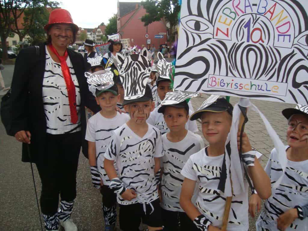 Eine Gruppe von Kindern und ein Erwachsener in Zebrakostümen stehen gemeinsam im Freien und halten ein buntes Schild mit der Aufschrift „ZEBRA-BANDE“ und „Bibrisschule“. Die Kinder haben bemalte Gesichter und Papierzebraohren.