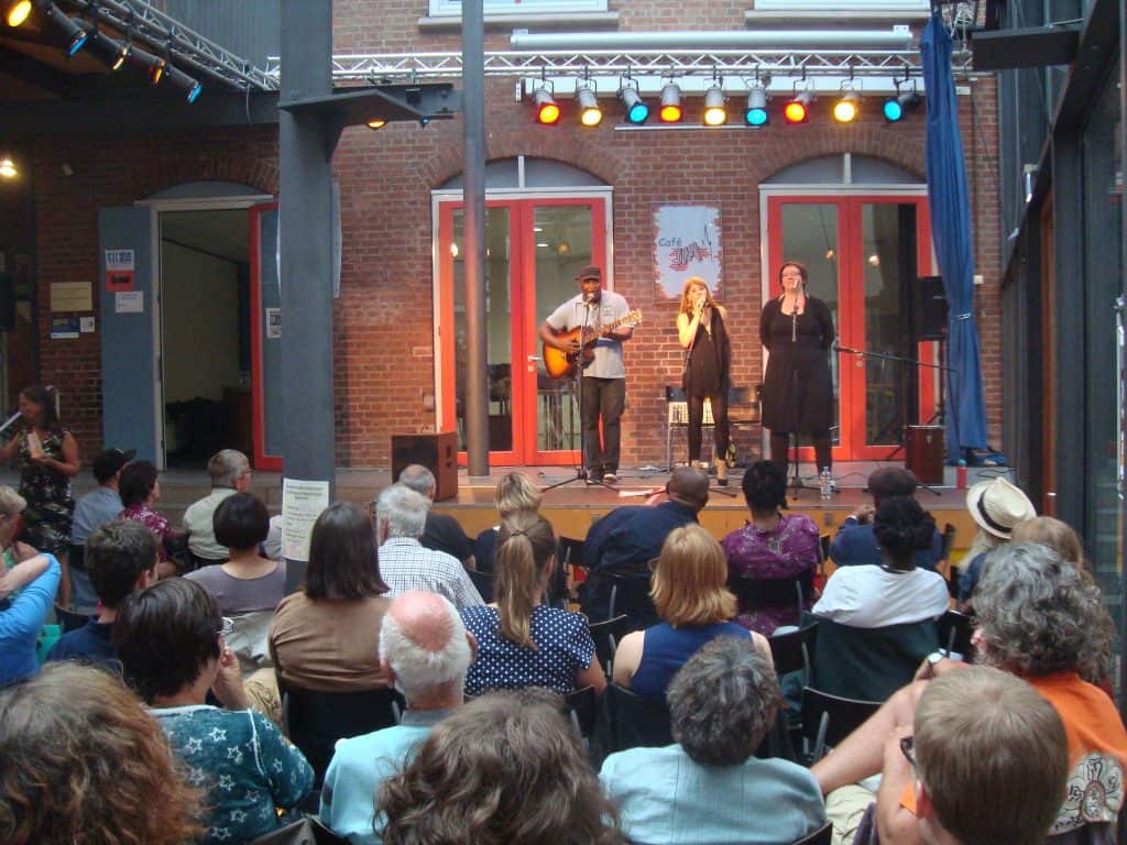 Eine kleine Band mit einem Gitarristen und zwei Sängern tritt auf einer bunt beleuchteten Bühne auf, während das Publikum aufmerksam zusieht. Im Hintergrund sind große Fenster und Backsteinmauern zu sehen.