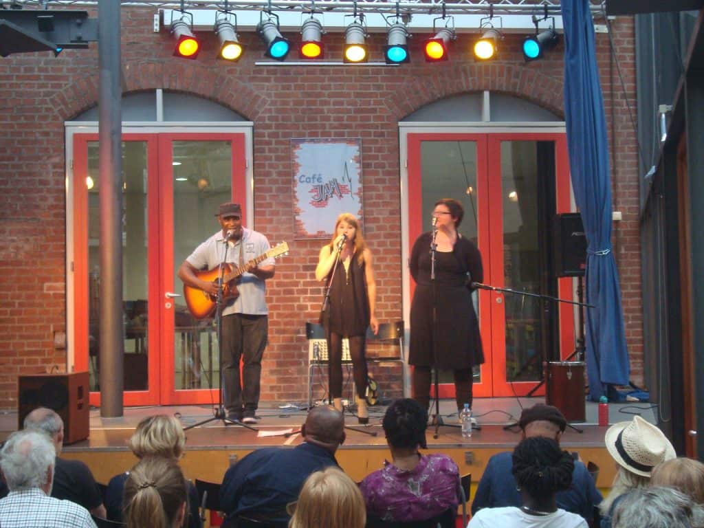 Drei Musiker treten auf einer kleinen Bühne im Innenbereich auf: Ein Mann spielt Gitarre, während zwei Frauen in Mikrofone singen. Sie blicken auf ein sitzendes Publikum, über ihnen bunte Lichter und hinter ihnen Ziegelwände.