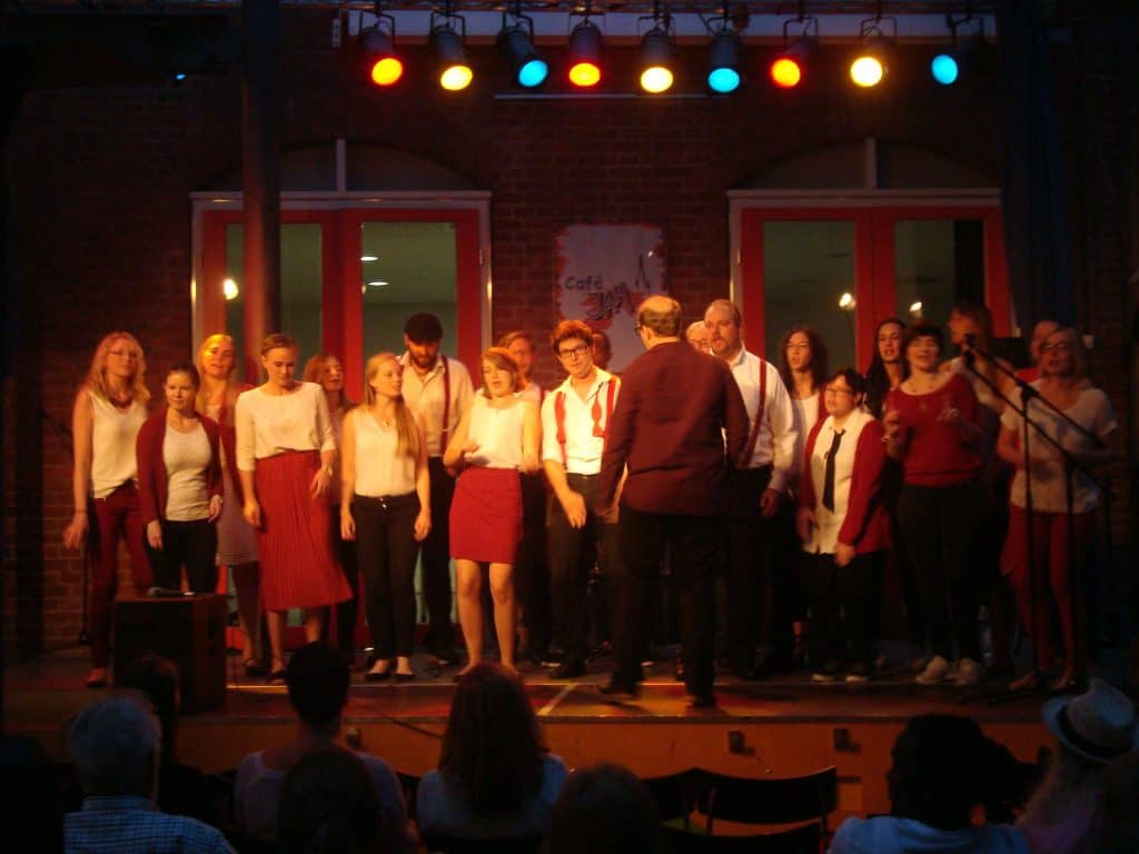 Ein Chor aus weiß-rot gekleideten Menschen tritt unter buntem Licht auf der Bühne auf. Der Dirigent steht der Gruppe gegenüber, und das Publikum schaut in den Sitzreihen im Vordergrund zu.